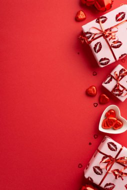 Sevgililer Günü konsepti. Üst görünümde hediye kutularının ambalaj kağıdında dikey fotoğrafı. Öpücük dudak desenli. Kalp şeklinde tabakta çikolata şekeri ve kopyalanmış kırmızı arka planda konfeti.