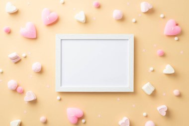 Sevgililer Günü konsepti. Beyaz fotoğraf çerçevesi, kalp şeklindeki şekerlemeler ve izole edilmiş bej arka planda yumuşak ponponlar.