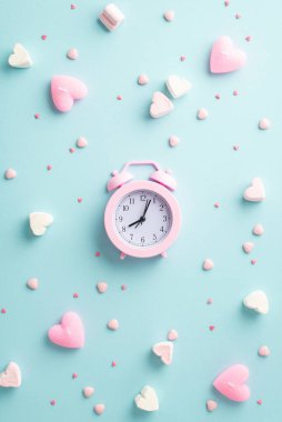 Sevgililer Günü konsepti. Pembe alarmlı saatin dikey resmi kalp şekerlemesi mumlar ve izole edilmiş pastel mavi arka plan üzerine serpiştirilmiş şekerlemeler.
