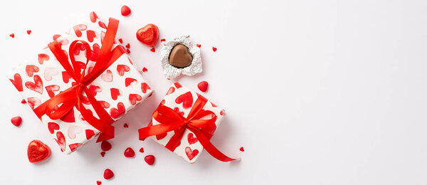 Valentine's Day concept. Top view photo of gift boxes with red ribbon bows heart shaped chocolate candies and sprinkles on isolated white background with copyspace