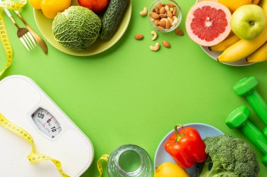 Uygun diyet konsepti. Meyve, sebze, çatal bıçak, halterli tabakların en üstteki fotoğrafı. Dambıllar ve teraziler izole edilmiş yeşil arkaplandalar. Ortasında da kopyalama alanı var.