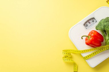 Slimming concept. Top view photo of bell pepper cauliflower and measuring tape wrapped around scales on isolated pastel yellow background with empty space