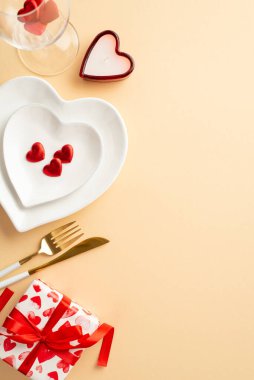 Valentine's Day concept. Top view vertical photo of heart shaped plates cutlery giftbox wineglass with confetti and candle on isolated pastel beige background with copyspace