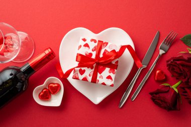 Saint Valentine's Day concept. Top view photo of heart shaped dish with present box cutlery wine bottle wineglass with confetti red roses and chocolate candies on isolated red background