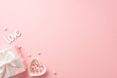 St Valentine's Day concept. Top view photo of giftbox with silk ribbon bow heart shaped dish with sprinkles and inscription love on isolated pastel pink background with copyspace