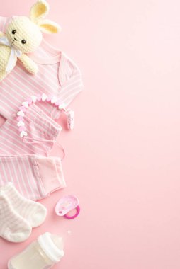Baby shower concept. Top view vertical photo of pink infant clothes shirt pants socks knitted bunny toy pacifier teether chain and milk bottle on isolated pastel pink background with copyspace
