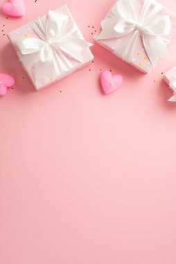 Valentine's Day concept. Top view vertical photo of gift boxes with white ribbon bows heart shaped candles and golden confetti on isolated pastel pink background with empty space