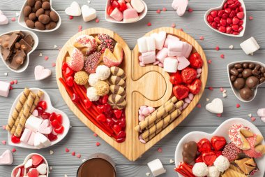 Valentine's Day concept. Top view photo of wooden heart shaped serving tray with confectionery plates jelly chocolate candies cookies and confetti on grey wooden table background