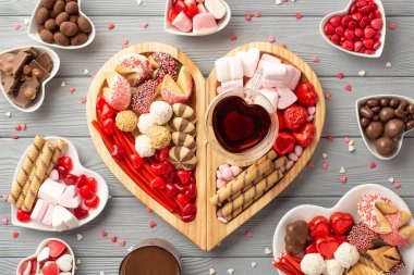 Valentine's Day concept. Top view photo of wooden heart shaped serving tray with sweets saucers candies cookies glass cup of drinking and confetti on grey wooden table background