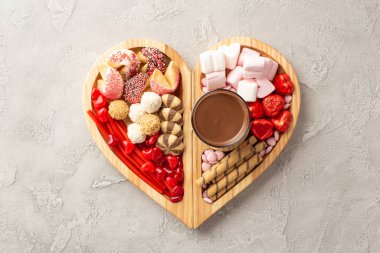 Valentine's Day concept. Top view photo of wooden heart shaped serving tray with sweets chocolate jelly candies cookies and glass cup of hot chocolate on concrete texture background