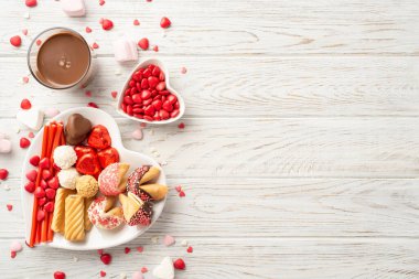 Valentine's Day celebration concept. Top view photo of heart shaped saucers with chocolate jelly candies cookies and glass of hot chocolate on white wooden desk background with copyspace