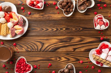 Valentine's Day concept. Top view photo of heart shaped saucers with sweets chocolate jelly candies cookies and glass of drinking on wooden desk background with copyspace in the middle