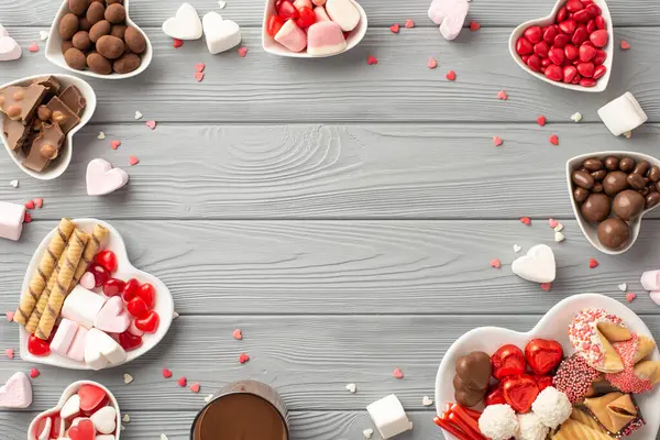 Sevgililer Günü konsepti. Kalp şeklindeki çay tabakları, çikolatalı kurabiyeler, bir bardak içki ve konfeti, gri tahta masa arka planında ve ortada boş bir alan.