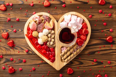 Valentine's Day concept. Top view photo of wooden heart shaped serving tray with sweets candies cookies and glass cup of drinking on wooden table background