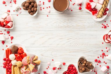 Valentine's Day concept. Top view photo of heart shaped crockery with chocolate jelly candies cookies and glass cup of hot chocolate on white wooden desk background with empty space in the middle