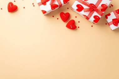 Valentine's Day concept. Top view photo of white gift boxes with ribbon bows red heart shaped candles and sprinkles on isolated pastel beige background with empty space
