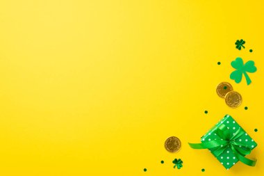 Saint Patrick's Day concept. Top view photo of small green present box with ribbon bow gold coins clover and trefoil shaped confetti on isolated yellow background with empty space