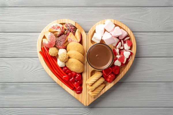 Valentine's Day concept. Top view photo of wooden heart shaped serving tray with confectionery candies cookies and glass cup of hot chocolate on grey wooden desk background
