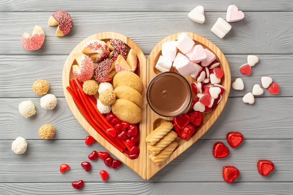 Valentine's Day concept. Top view photo of wooden heart shaped serving tray with confectionery candies cookies and glass cup of hot chocolate on grey wooden desk background