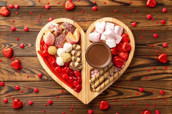 Valentine's Day celebration concept. Top view photo of wooden heart shaped serving tray with confectionery candies cookies and glass of hot chocolate on wooden desk background