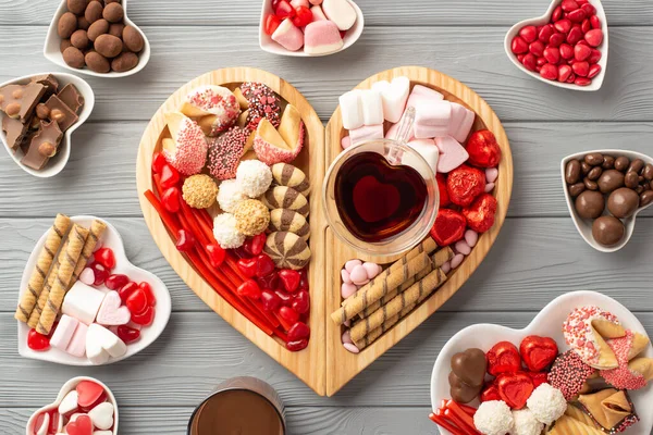 Valentine's Day concept. Top view photo of wooden heart shaped serving tray with sweets plates chocolate jelly candies cookies and glass cup of drinking on grey wooden desk background