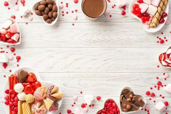 Valentine's Day concept. Top view photo of heart shaped crockery with chocolate jelly candies cookies and glass cup of hot chocolate on white wooden desk background with empty space in the middle
