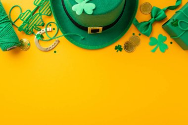 Top view photo of leprechaun cap present box clover shaped party glasses spool of twine bow-tie gold coins shamrocks horseshoe and confetti on isolated yellow background with empty space clipart