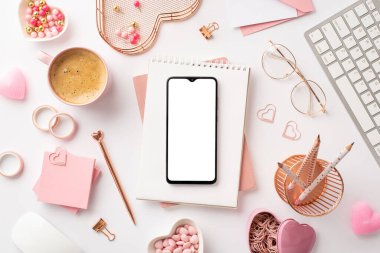 Valentine's Day concept. Top view photo of smartphone over notepads keyboard glasses stationery holders saucer with sprinkle candles and cup of coffee on isolated white background with blank space