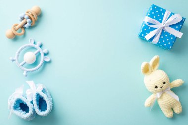Baby shower concept. Top view photo of blue knitted booties present box with bow bunny toy teether and wooden rattle on isolated pastel blue background with copyspace