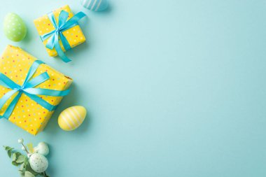 Easter decor concept. Top view photo of bright yellow gift boxes with blue ribbon bows and bouquet with colorful easter eggs on isolated pastel blue background with empty space