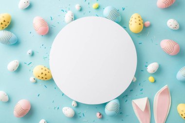 Easter decorations concept. Top view photo of white circle colorful easter eggs bunny ears and sprinkles on isolated light blue background with copyspace