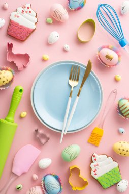 Easter cooking concept. Top view vertical photo of blue plates cutlery kitchen utensils colorful easter eggs in paper baking molds and gingerbread on isolated pastel pink background