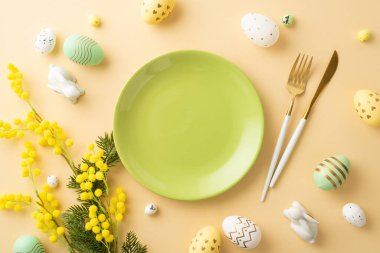 Easter celebration concept. Top view photo of empty green plate knife fork colorful easter eggs ceramic easter bunnies and mimosa flowers on isolated beige background with copyspace