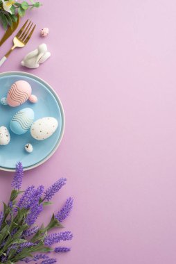 Easter celebration concept. Top view vertical photo of blue plate with easter eggs cutlery ceramic rabbit and bunch of lavender flowers on isolated lilac background with empty space