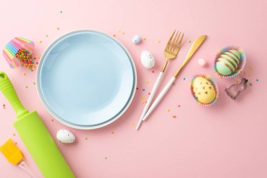 Easter celebration concept. Top view photo of empty blue plates knife fork rolling pin brush colorful easter eggs in paper baking molds and sprinkles on isolated pastel pink background