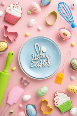Top view vertical photo of blue plate with inscription happy easter kitchen utensils colorful easter eggs in paper baking molds and cake shaped gingerbread on isolated pastel pink background