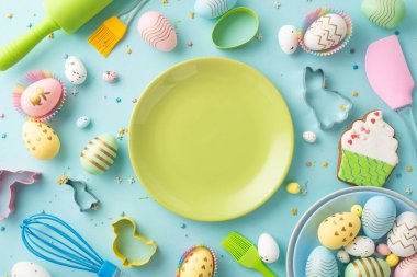 Easter celebration concept. Top view photo of green dish colorful eggs rolling-pin whisk brush silicone spatula baking molds gingerbread and sprinkles on isolated pastel blue background with copyspace