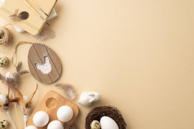 Easter decorations concept. Top view photo of easter eggs in wooden holder quail nest ceramic rabbit sketchbook paintbrushes and feathers on isolated pastel beige background with copyspace