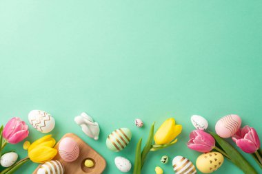 Easter celebration concept. Top view photo of colorful easter eggs yellow and pink tulips ceramic bunny and wooden egg holder on isolated turquoise background with copyspace