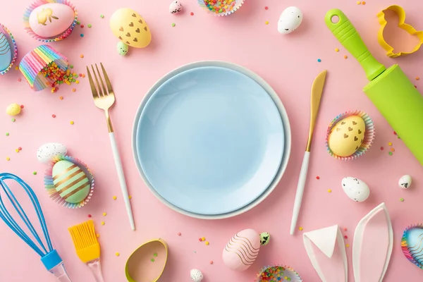 Easter celebration concept. Top view photo of empty blue dishes knife fork rolling pin whisk colorful easter eggs in paper baking molds bunny ears and sprinkles on isolated pastel pink background