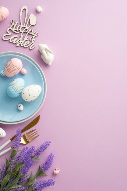 Easter concept. Top view vertical photo of blue dish with easter eggs knife fork inscription happy easter ceramic bunny and bouquet of lavender flowers on isolated lilac background