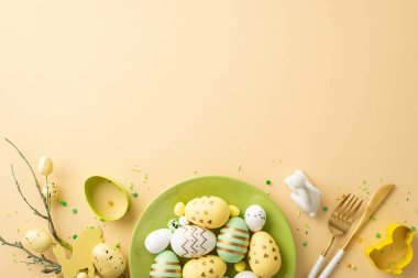 Easter celebration concept. Top view photo of green plate with colorful easter eggs cutlery ceramic easter bunny baking molds easter bouquet and sprinkles on isolated beige background with copyspace