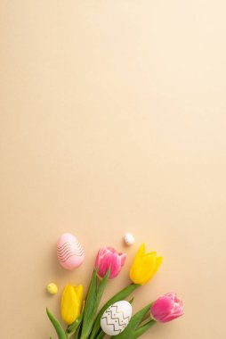 Easter decor concept. Top view vertical photo of bouquet of yellow and pink tulips and colorful easter eggs on isolated pastel beige background with copyspace