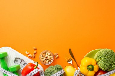 Zayıflatıcı konsept. Dambıl ve sebzeli mezura ölçü tabağı, kabuklu çan biberi, lahana ve çatal bıçak takımlarının izole edilmiş turuncu arka planda fotoğrafı.