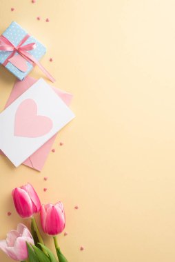 Anneler Günü kutlaması konsepti. Pembe lalelerin dikey üst görüntüsü küçük mavi kutu zarfı ve izole edilmiş pastel bej arkaplan üzerine kalp şeklinde serpiştirilmiş boş alan.