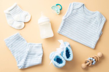 Bebek aksesuarları konsepti. Mavi gömlekli külotların üst görüntüsü patik örgü çoraplar süt şişesi sakinleştirici ve ahşap çıngırak izole edilmiş pastel bej arka plan