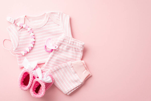 Baby accessories concept. Top view photo of pink infant clothes shirt pants small knitted booties pacifier and teether chain on isolated pastel pink background with copyspace