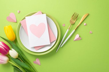 Anneler Günü konsepti asgari düzeyde. Tabaklar, çatal bıçak takımı, laleler, tabakta tebrik kartı ve açık yeşil arka planda dekoratif kalpler.