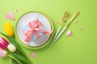 Annemin masa hazırlama konsepti. Tabaklar, çatal bıçak takımı, laleler, tabakta ipek kurdele ve açık yeşil arka planda dekoratif kalplerden oluşan bir hediye kutusu. Minimalist ve zarif tasarım