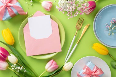 Anneler Günü masa konsepti. Renkli tabakların üst görünümü düz, açık zarf içinde açık kağıt kart ileti, çatal bıçak takımı ve açık yeşil zemin üzerinde laleler için